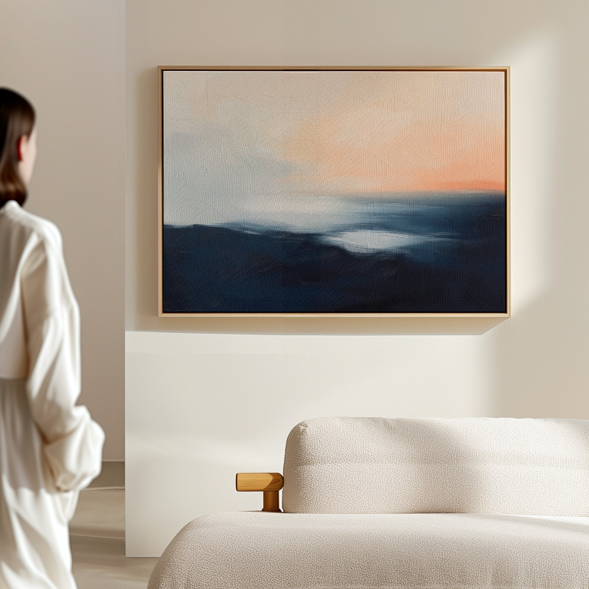 A person stands in a softly lit room, looking at an abstract painting with blue and peach tones hanging above a light-colored sofa. The scene is minimalist and calm.