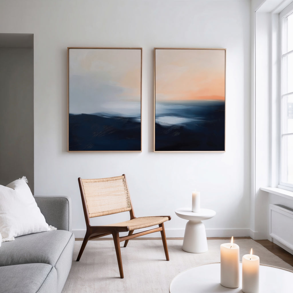 A modern living room with a gray sofa, wooden chair, and small white table holding a lit candle. Abstract blue and orange landscape wall art, the Dusk Ridge Set of 2, hangs on a white wall. More candles are on a nearby table.