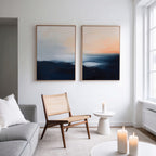 A modern living room with a gray sofa, wooden chair, and small white table holding a lit candle. Abstract blue and orange landscape wall art, the Dusk Ridge Set of 2, hangs on a white wall. More candles are on a nearby table.