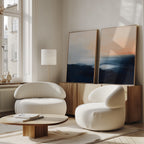 A modern living room features the Dusk Ridge Set of 2 white curved armchairs, a low wooden coffee table, and a sideboard with serene wall art. Sunlight pours through large windows, creating a warm and inviting atmosphere.