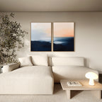 A modern living room with a cream sofa, a small wooden coffee table, a large leafy plant, and the Dusk Ridge Set of 2—abstract landscape paintings in blue and peach tones—adds a calming touch to the space.