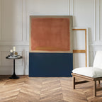 A modern minimalist room with herringbone wood floors features the Dusk Stripe Abstract Canvas Art—rust on top, dark blue below—leaning against a white paneled wall beside an empty frame, black table, and white chair.