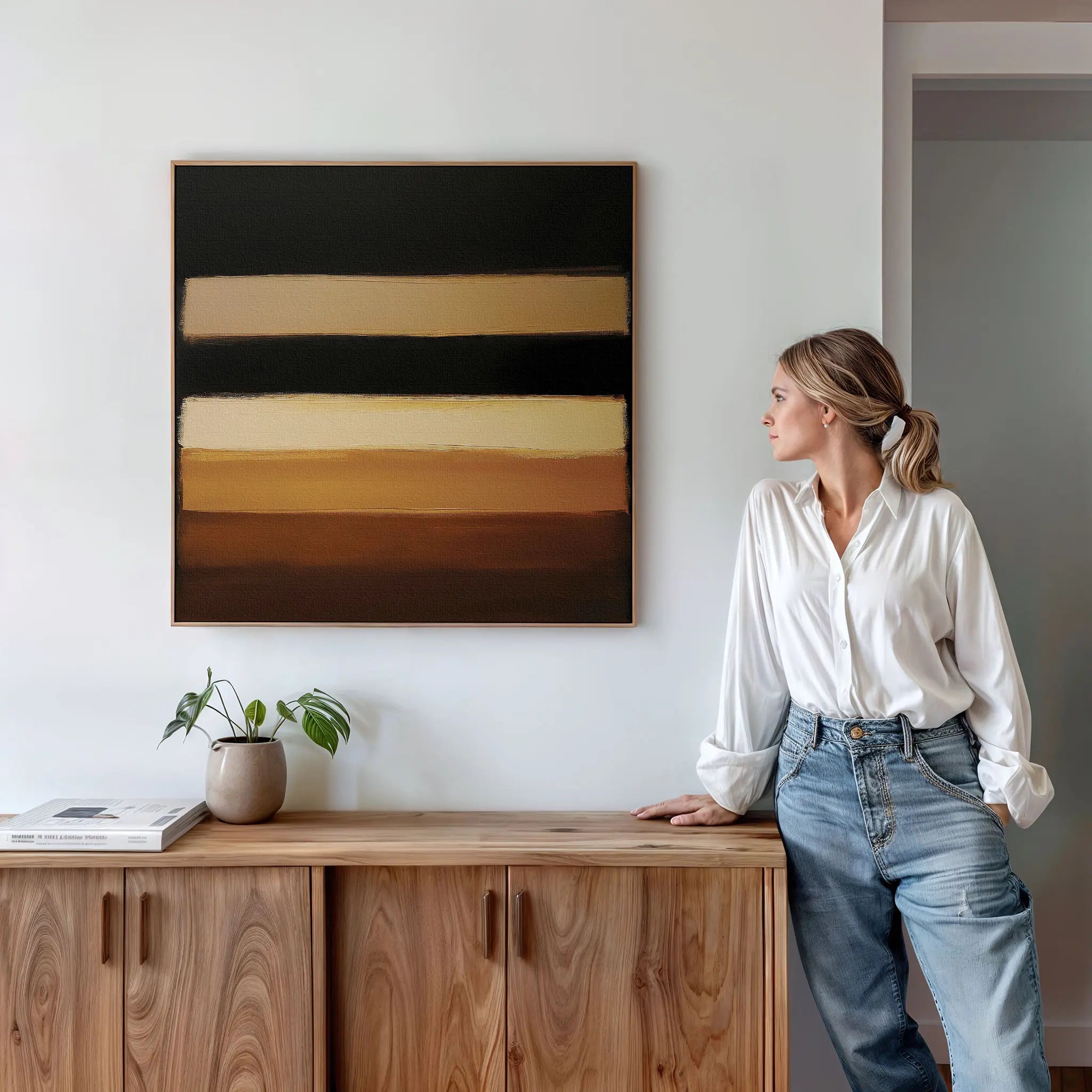 A woman in a white blouse and jeans stands by a wooden cabinet, admiring the Earth Layers Canvas Art with black, beige, and brown stripes on a white wall. A plant and book on the cabinet add to the serene living room vibe.