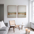 A minimal, modern living room features a woven chair, round white side table, lit candles, gray sofa with white pillow, and the Earthly Hues Set of 2 as minimalist wall art on a white wall by large windows.