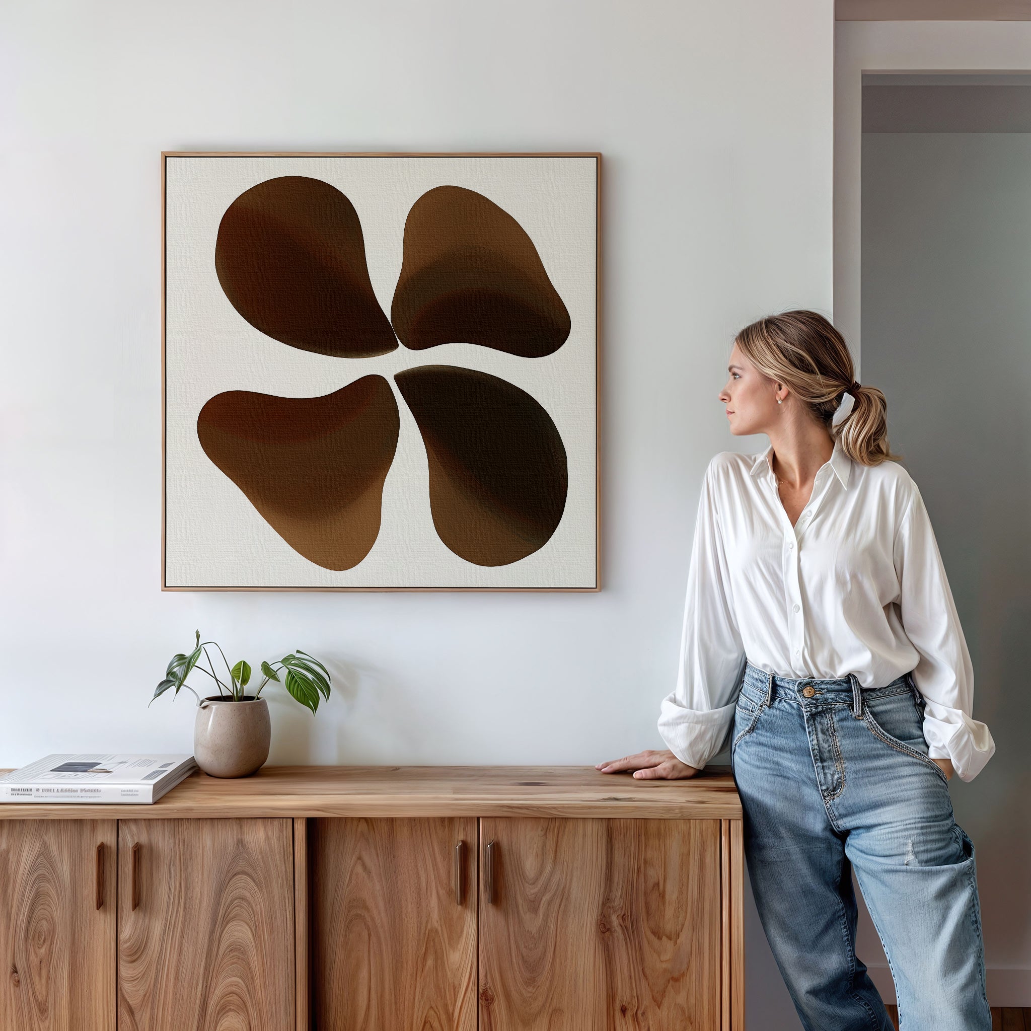 A woman in a white shirt and jeans leans against a wooden cabinet, gazing at the Earthy Blooms Canvas Art with its four rounded shapes above her. A plant and book on the cabinet enhance the serene, minimalist scene.