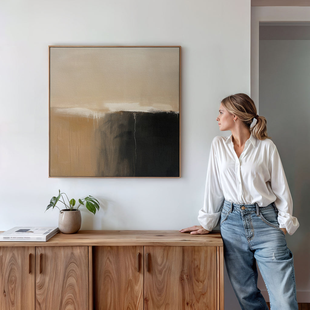 A woman in a white blouse and jeans leans on a wooden cabinet, admiring the Earthy Cliffs Abstract Canvas Art in brown, beige, and black tones above. A plant and books decorate the living room cabinet.
