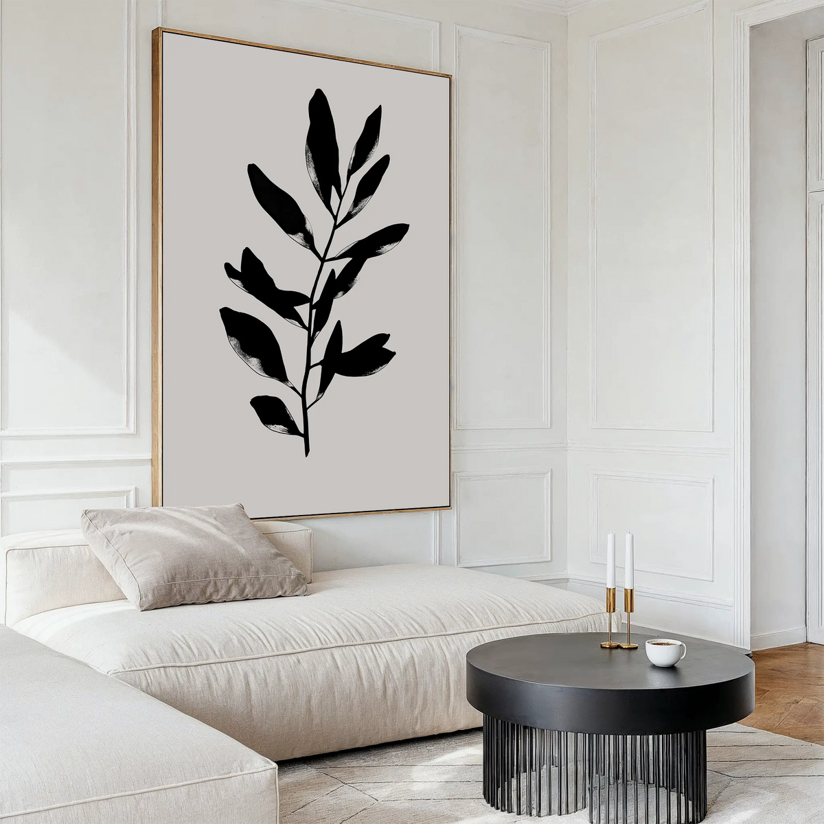 A modern living room features a cream sectional sofa, light pillows, a round black coffee table, and the Ebony Leaf Canvas Art. The neutral decor includes white paneled walls, adding minimalist style throughout the space.