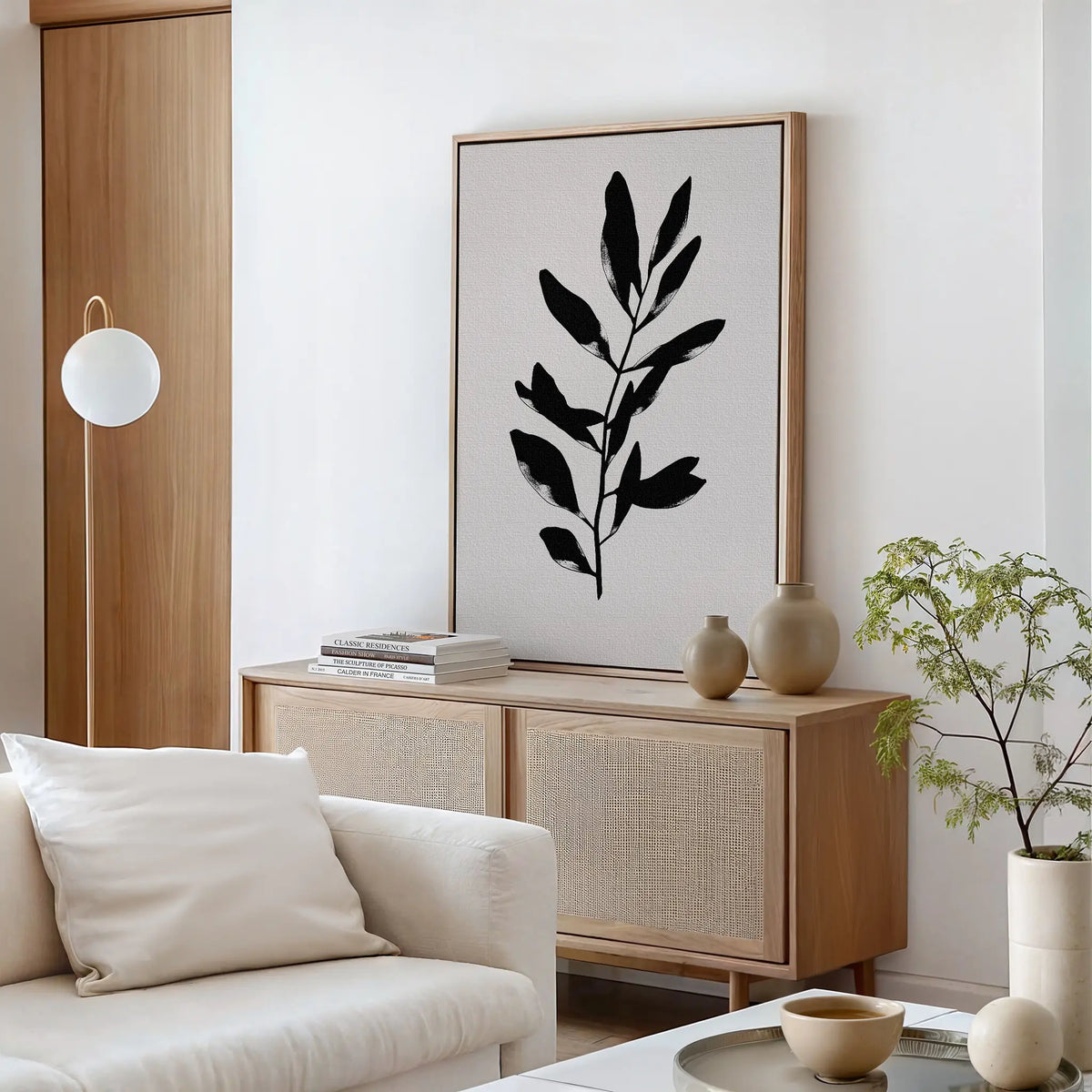 A minimalist living room featuring a light wood sideboard, Ebony Leaf Canvas Art, neutral pottery, a small potted plant, and a white sofa with a plush cushion—perfect for modern interiors.