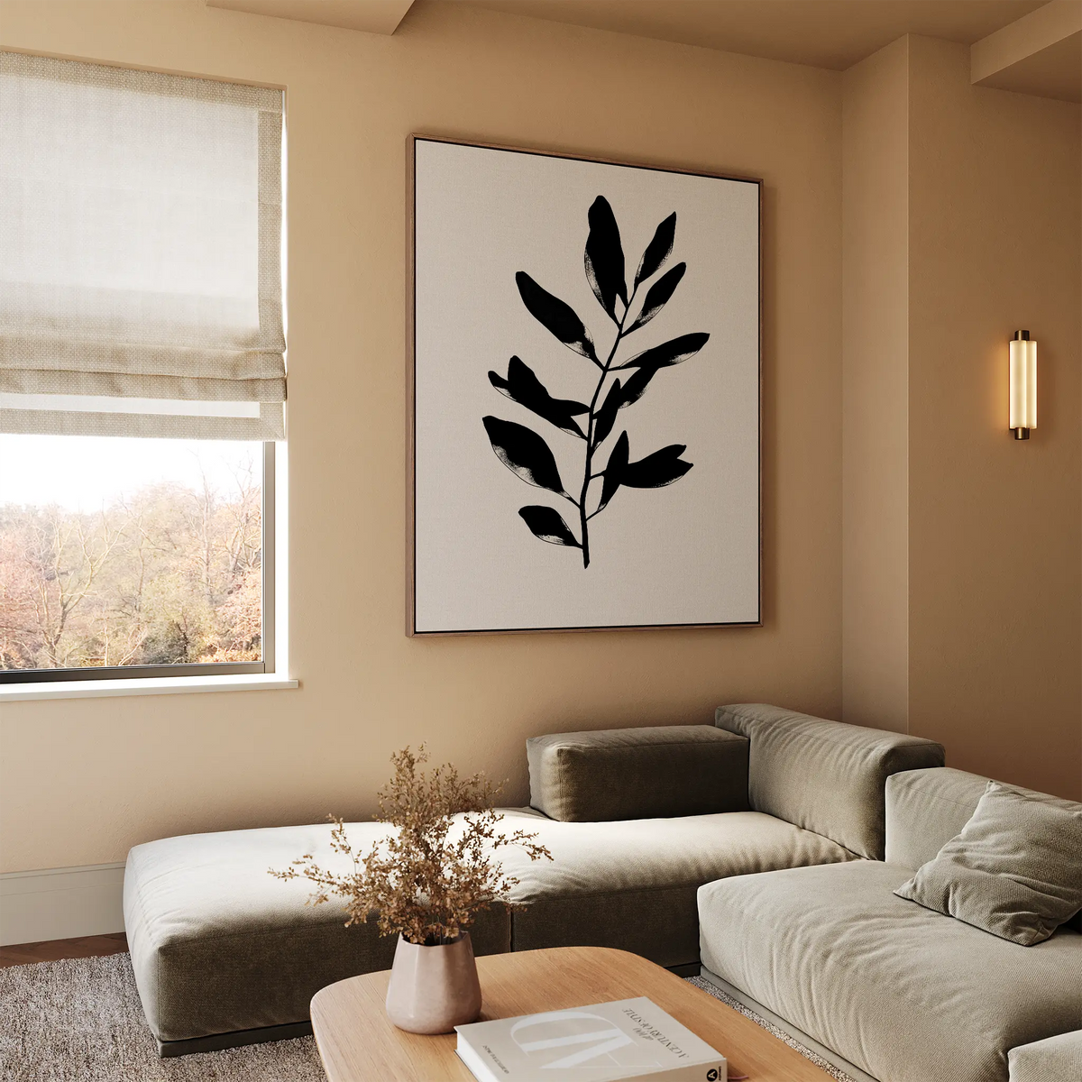 A modern living room with neutral tones features the Ebony Leaf Canvas Art—a large abstract black leaf—paired with a beige sectional, wooden coffee table with dried flowers, and a roman shade, perfect for stylish contemporary interiors.