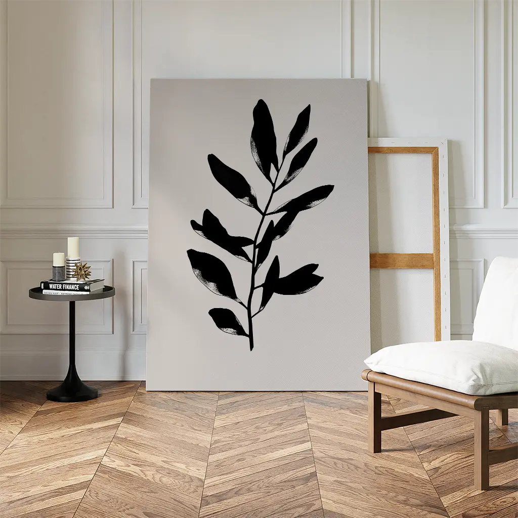 A minimalist room with herringbone wood floors features Ebony Leaf Canvas Art—its black leaf silhouette propped against the wall next to an empty frame. A small round table with candles and a white chair complete the modern interior.
