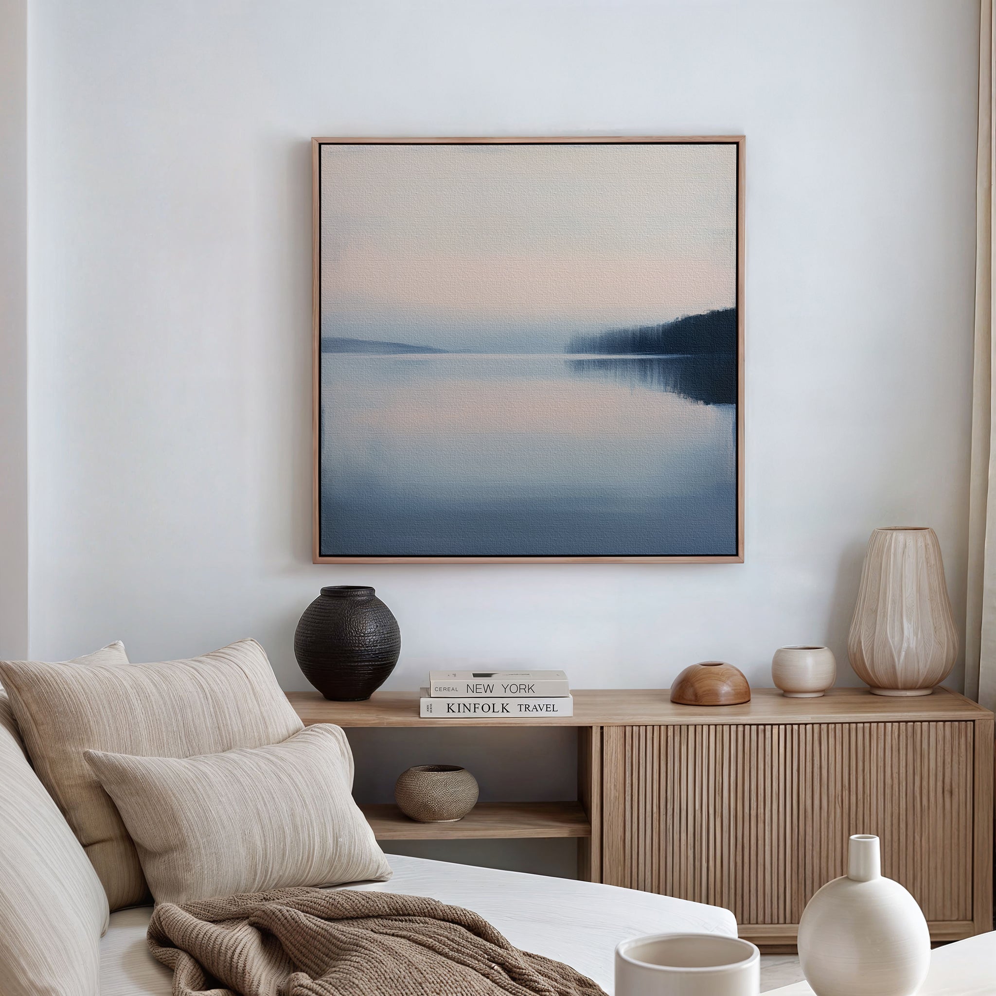 A serene, minimalist living room features a beige sofa, textured cushions, and a wooden sideboard with books and pottery. Above it hangs Echo Lake Canvas Art—a tranquil landscape—adding calm to the white wall.