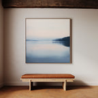 A minimalist room features a wooden bench with a brown leather cushion below Echo Lake Canvas Art, a large framed lake scene with soft blue and pink tones, hanging on a light-colored wall.