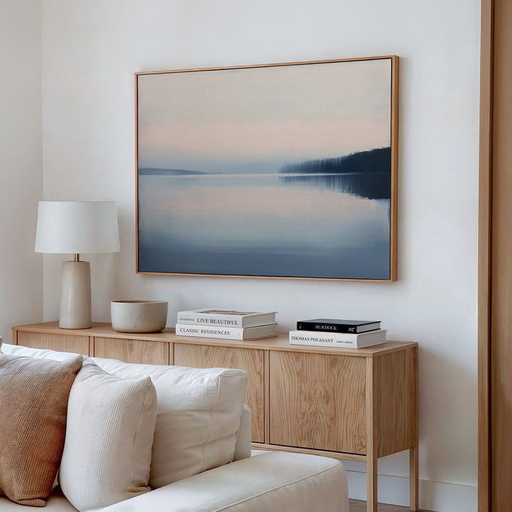 A minimal living room features a light wood sideboard with neutral decor, stacked books, a white lamp, and bowl. Above hangs the Echo Lake Canvas Art – Horizontal, depicting a calm lake at sunset. A cream sofa is partially visible in the foreground.