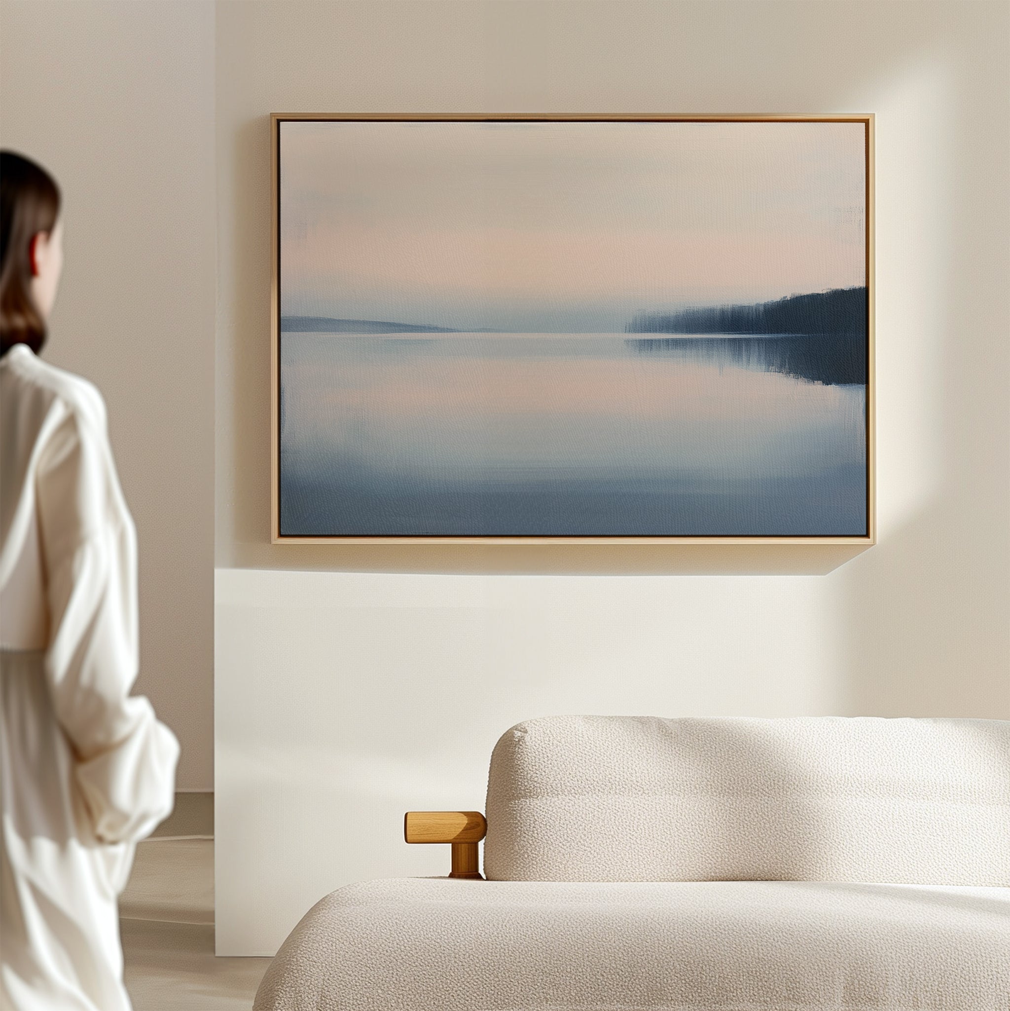 A person in a white shirt stands in a minimal, light-filled room with a beige sofa and serene wall art—a large, framed Echo Lake Canvas Art – Horizontal featuring soft blue and pink tones—adding calming bedroom decor.