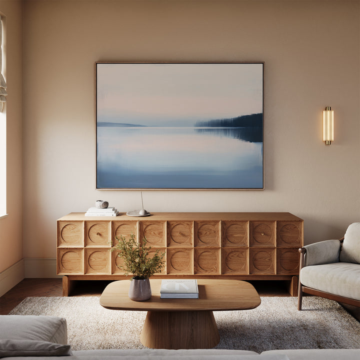 A modern living room featuring a wooden coffee table, textured sideboard with decor and lamp, neutral sofa, beige rug, and serene Echo Lake Canvas Art – Horizontal—a large framed landscape of a misty lake—on the wall.