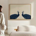 A person in a white shirt admires the Elegant Plumage Canvas Art—two blue peacocks face to face—displayed above a beige couch, adding charm and sophistication to the softly lit, minimalist living room decor.