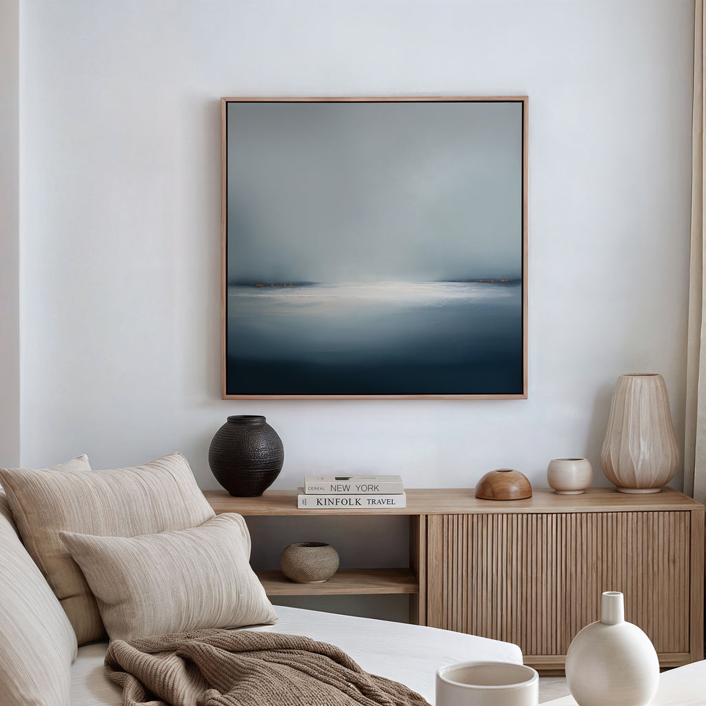 A cozy living room featuring a beige sofa with neutral pillows and throws, a wooden sideboard with books, vases, decor accents, and minimalist wall art—the Emberline Stillness Canvas Art—displayed on a white wall.