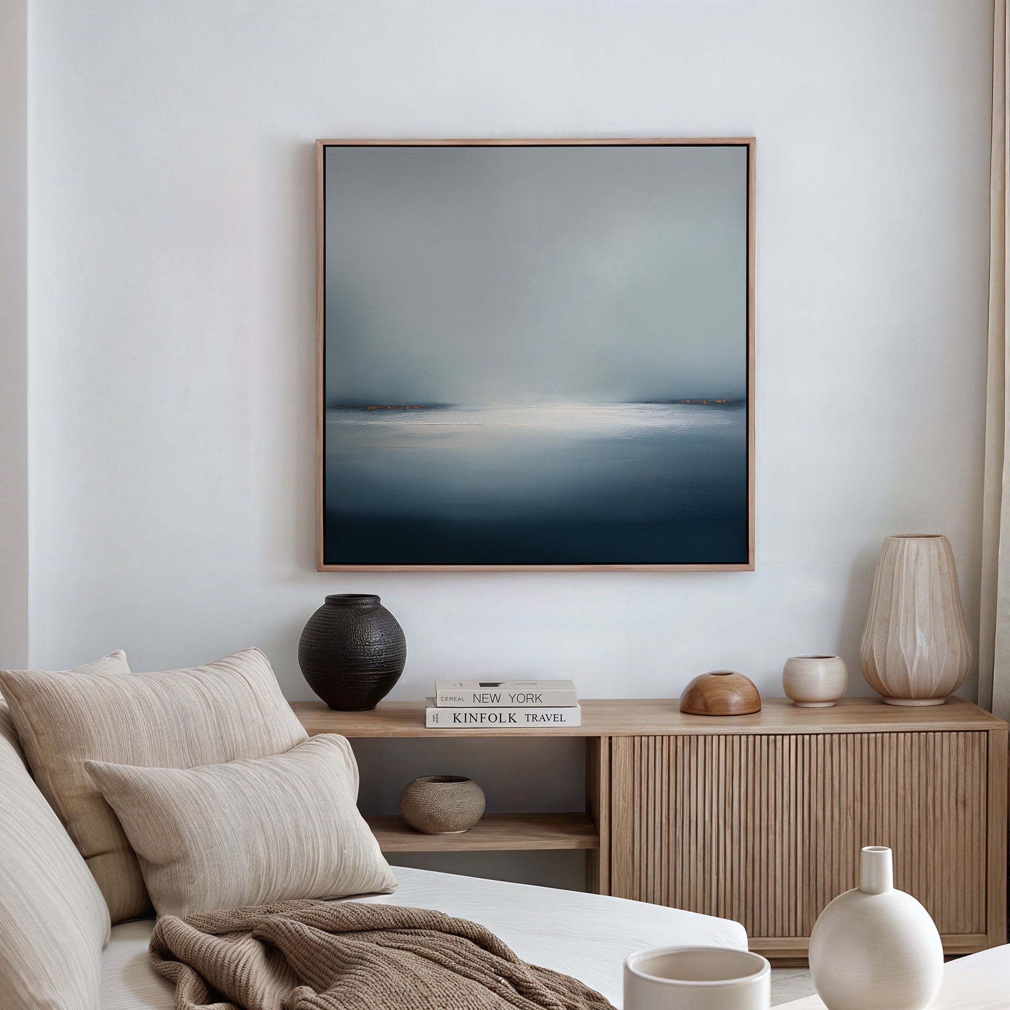 A cozy living room featuring a beige sofa with neutral pillows and throws, a wooden sideboard with books, vases, decor accents, and minimalist wall art—the Emberline Stillness Canvas Art—displayed on a white wall.