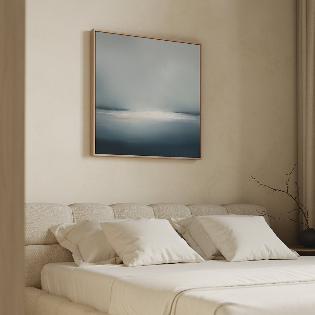 A minimal bedroom features a beige upholstered bed with white bedding, soft lighting, and the Emberline Stillness Canvas Art in blue and gray above the headboard. A bare branch on the side table completes the modern look.
