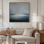 A modern living room features a beige sofa, cream cushions, a side table with lamp, and dried flowers in a vase. The space is elevated by minimalist décor including the Emberline Stillness Canvas Art—a large abstract blue and white painting.
