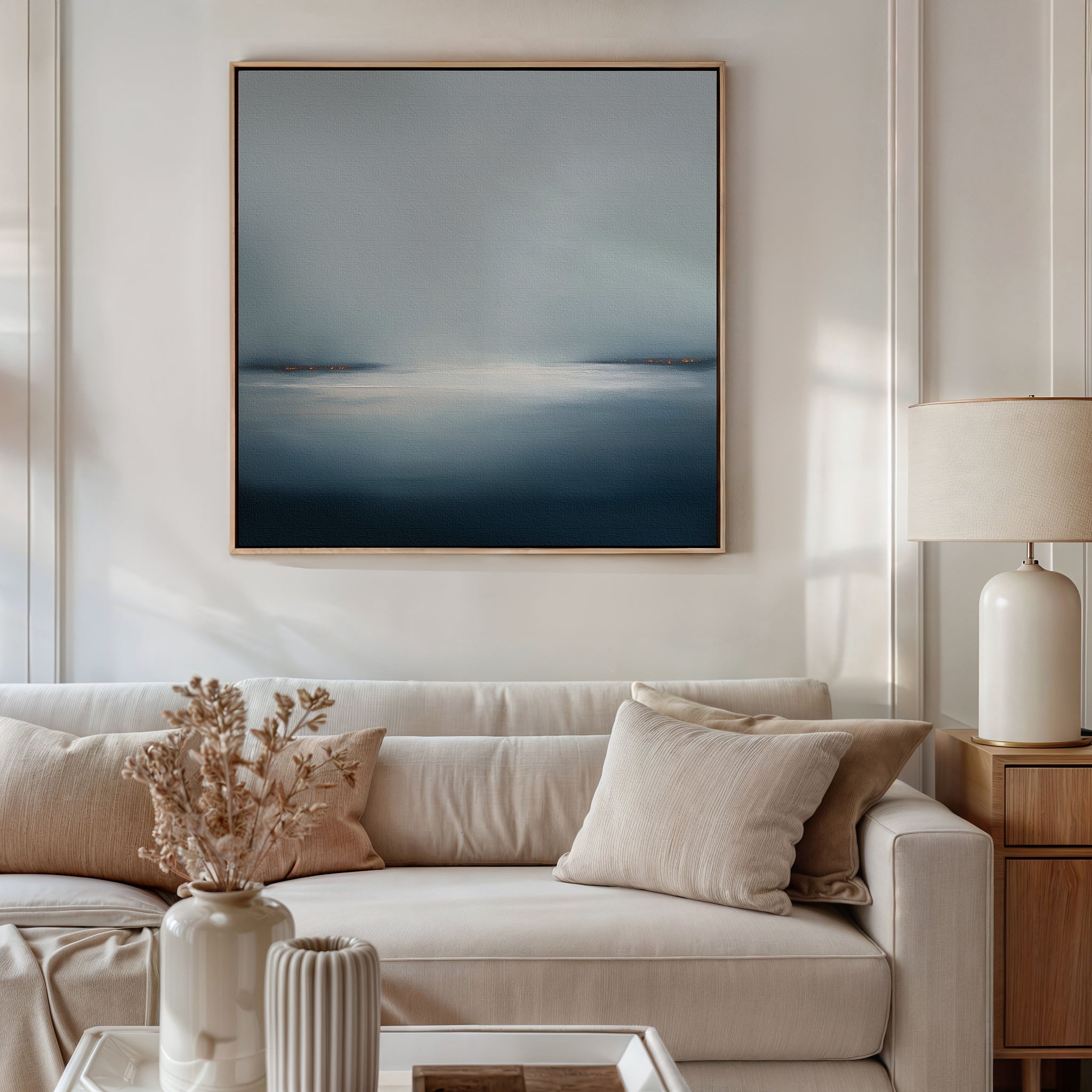 A modern living room features a beige sofa, cream cushions, a side table with lamp, and dried flowers in a vase. The space is elevated by minimalist décor including the Emberline Stillness Canvas Art—a large abstract blue and white painting.