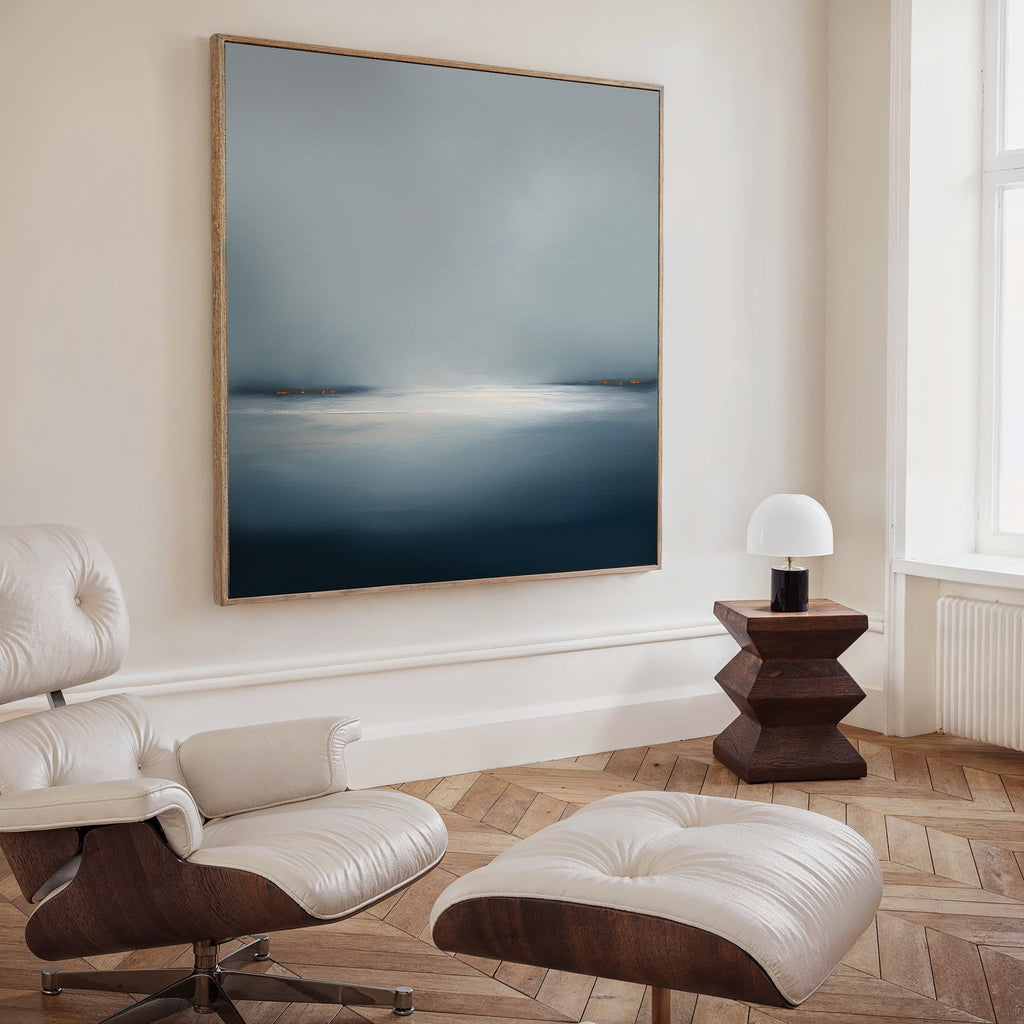 A modern, minimalist living room featuring a white lounge chair and ottoman, wooden herringbone floor, geometric side table, lamp, and the Emberline Stillness Canvas Art in blue and gray—perfect inspiration for contemporary interiors.