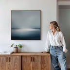 A woman in a white blouse and jeans leans against a wooden cabinet, admiring the Emberline Stillness Canvas Art with blue and white tones. A potted plant and books enhance the modern space, blending style and comfort.