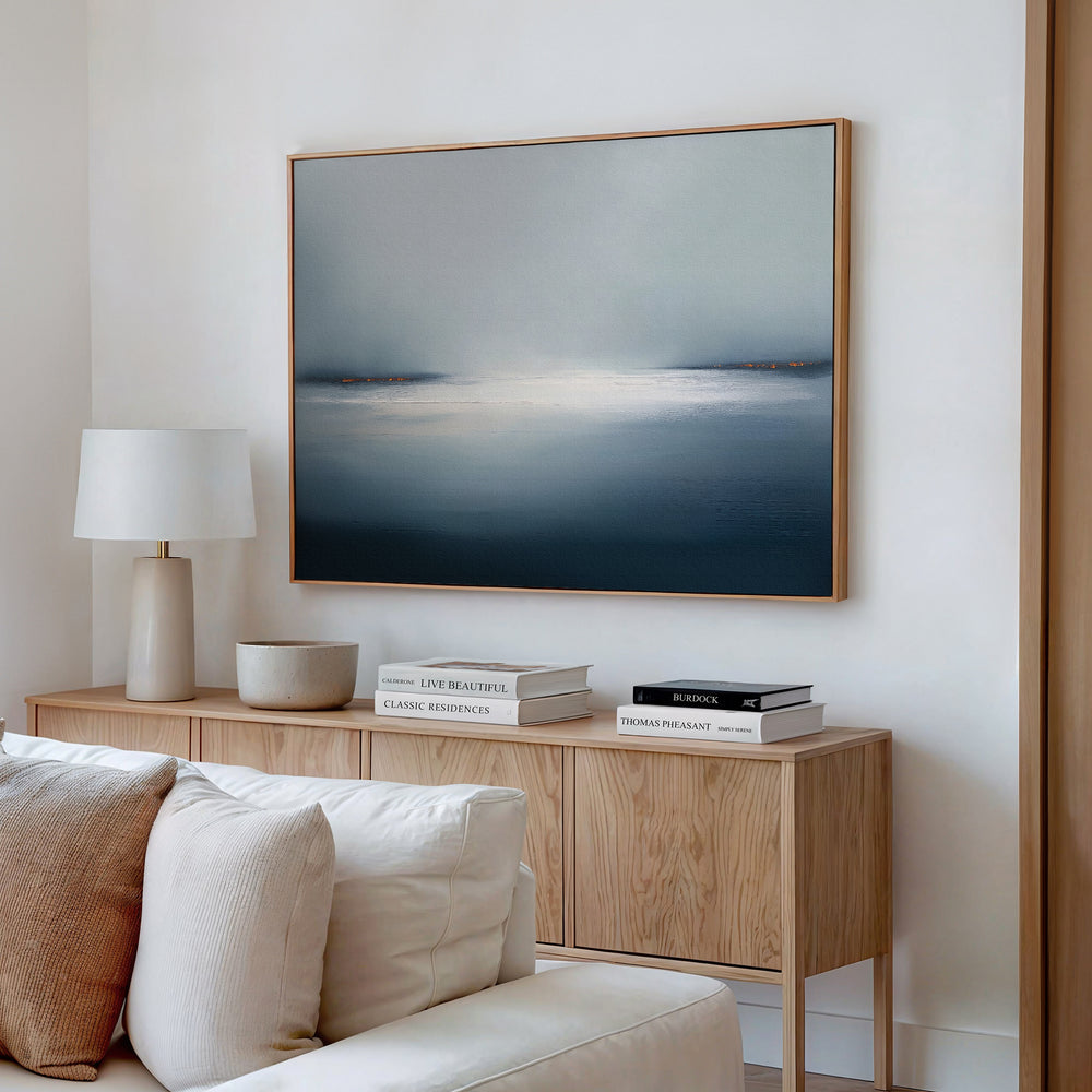 A minimalist living room featuring a light wood sideboard, white lamp, stacked books, beige bowl, white couch, and large wall art—the Emberline Stillness Canvas Art – Horizontal with soft blue and white tones—displayed on the wall.