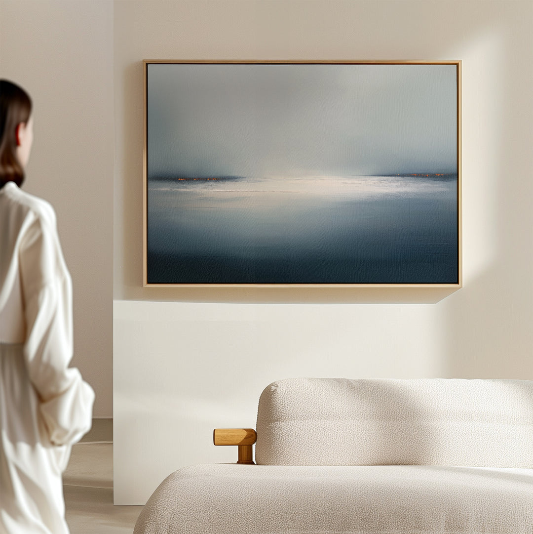 A person admires the Emberline Stillness Canvas Art – Horizontal, featuring blue and white tones above a light-colored sofa in a softly lit room. The serene artwork perfectly complements modern minimalist interiors.