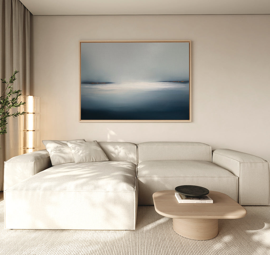 A modern living room with a light beige sectional sofa, a wooden coffee table with a black bowl, neutral walls, and the Emberline Stillness Canvas Art – Horizontal above the sofa is brightened by natural light filtering through curtains.