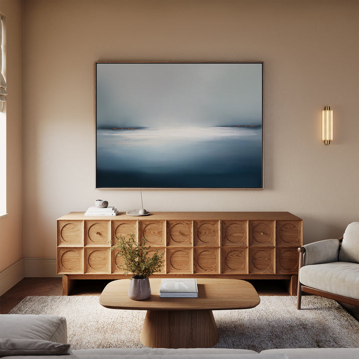 A modern living room with neutral tones features a wooden sideboard, a low coffee table, a light sofa, and the Emberline Stillness Canvas Art – Horizontal above, accented by a wall sconce and flowers—ideal for modern style fans.