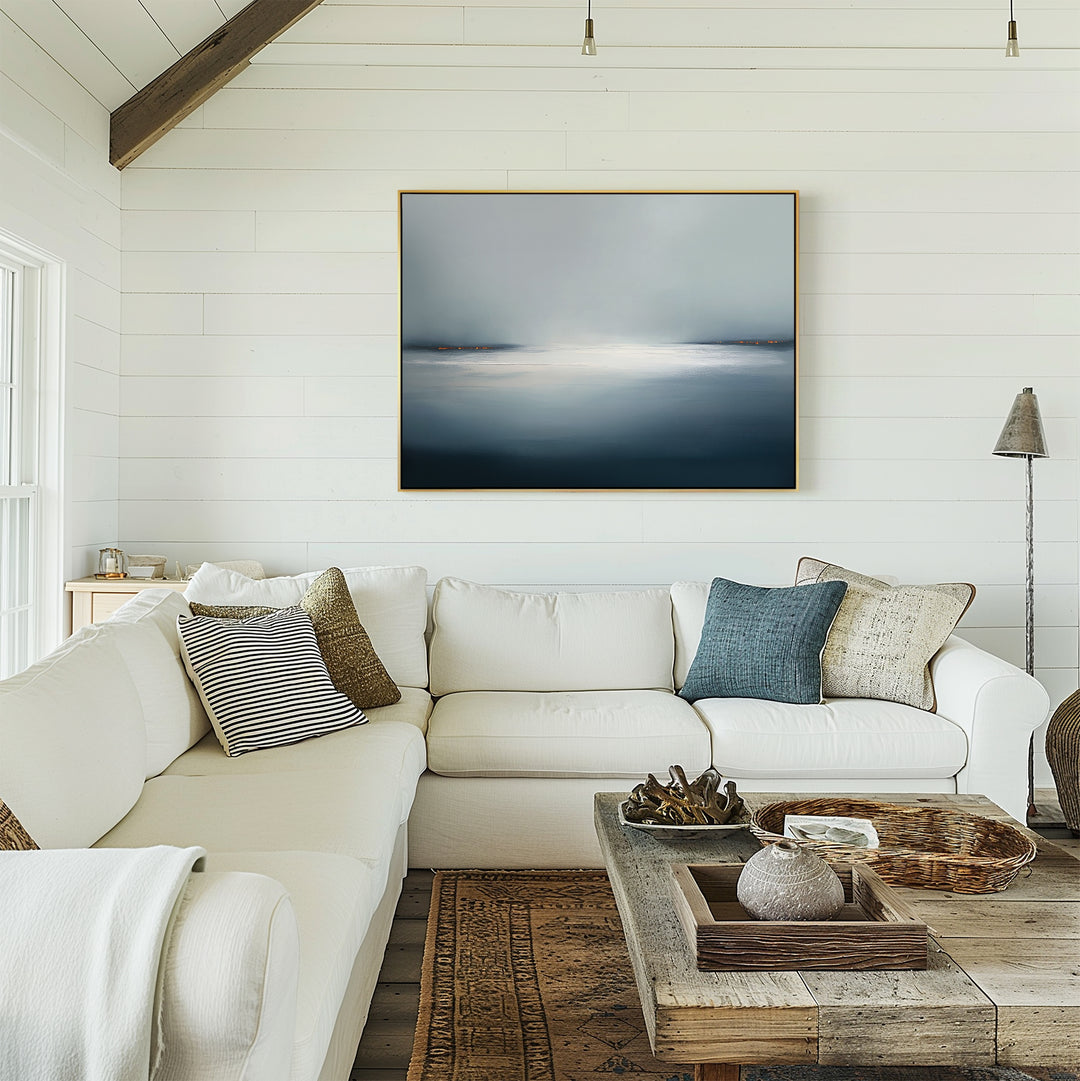 A bright, airy living room features a white sectional sofa, assorted pillows, a rustic wood coffee table, and the Emberline Stillness Canvas Art – Horizontal on a white shiplap wall. Natural light from the left window enhances the modern space.