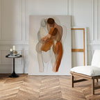 The Embrace Abstract Canvas Art, featuring two intertwined human figures with transparent shapes, leans against a white paneled wall. Nearby sit a chair, an empty frame, and a side table with candles and books for a minimalist look.