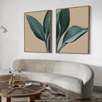 A modern living room with a curved beige sofa, a round wooden coffee table with an open book and vase, and the Emerald Poise Set of 2—large minimalist wall art prints with green leaves that elevate the decor.