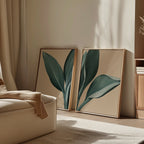 The Emerald Poise Set of 2 features framed minimalist wall art prints with green leaves, leaning against a beige wall in a sunlit room with a cream armchair and neutral decor for a calm, modern look.