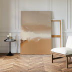 A minimalist room with herringbone wood floors features the Ethereal Plains Abstract Canvas Art in beige and brown tones, leaning against the wall beside an empty frame, a white cushioned chair, and a small black table with books and candles.