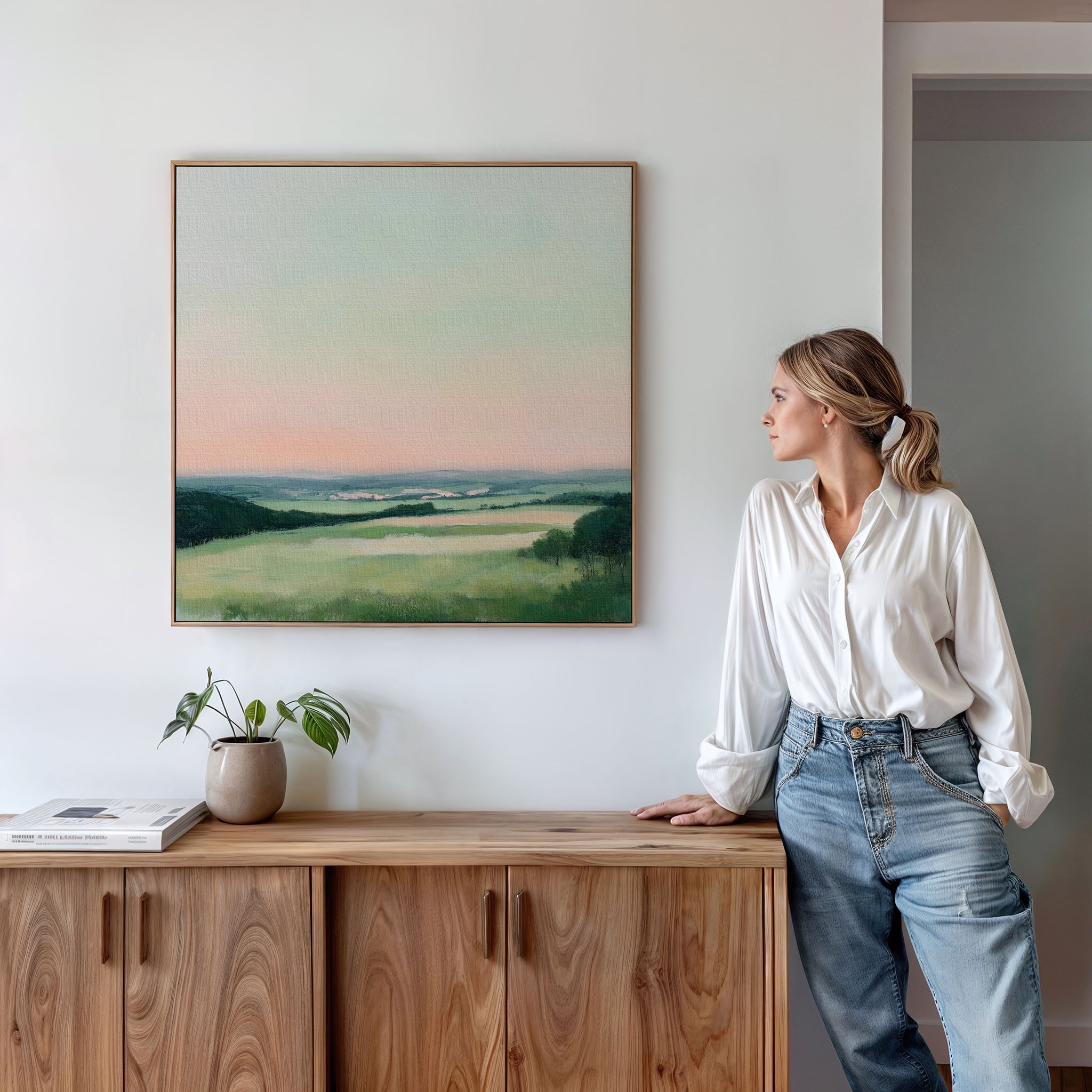 A woman in a white blouse and jeans leans against a wooden sideboard, admiring the Evening Hues Abstract Canvas Art, a striking landscape print and perfect living room decor, displayed on the white wall above her.