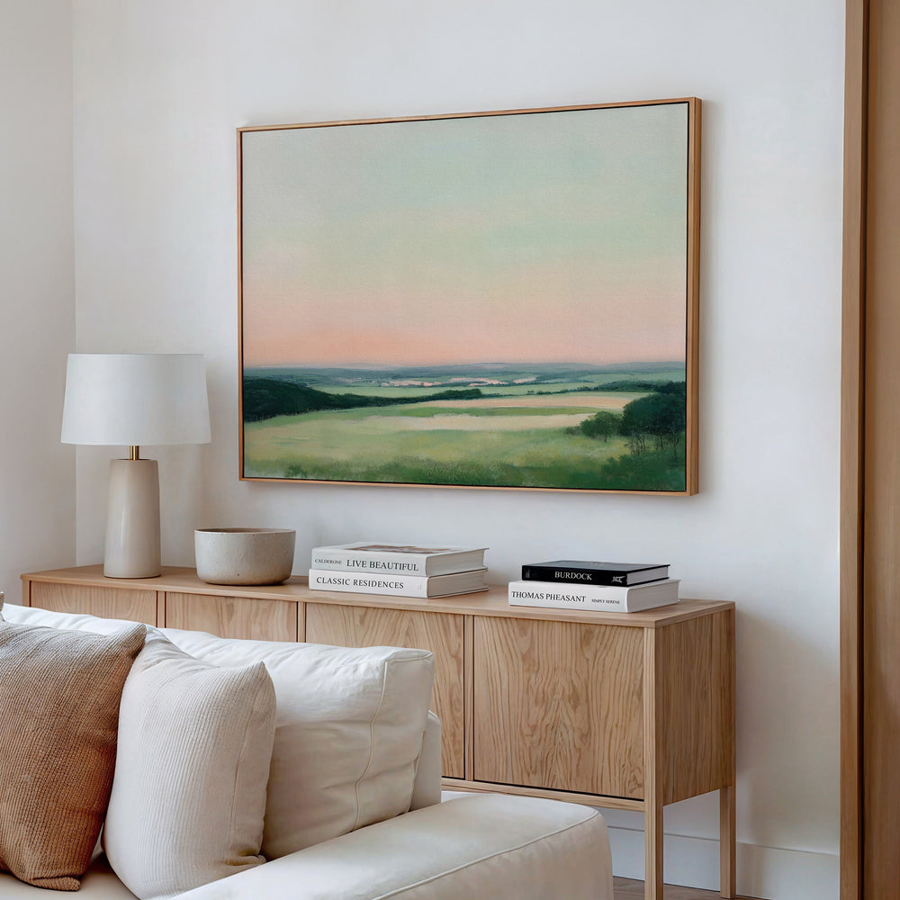 A modern living room features a light wooden console with decorative books, a lamp, and a bowl. Above it hangs the Evening Hues Canvas Art – Horizontal, depicting green fields under a pastel sky. A white sofa sits in the foreground.
