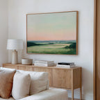 A modern living room features a light wooden console with decorative books, a lamp, and a bowl. Above it hangs the Evening Hues Canvas Art – Horizontal, depicting green fields under a pastel sky. A white sofa sits in the foreground.