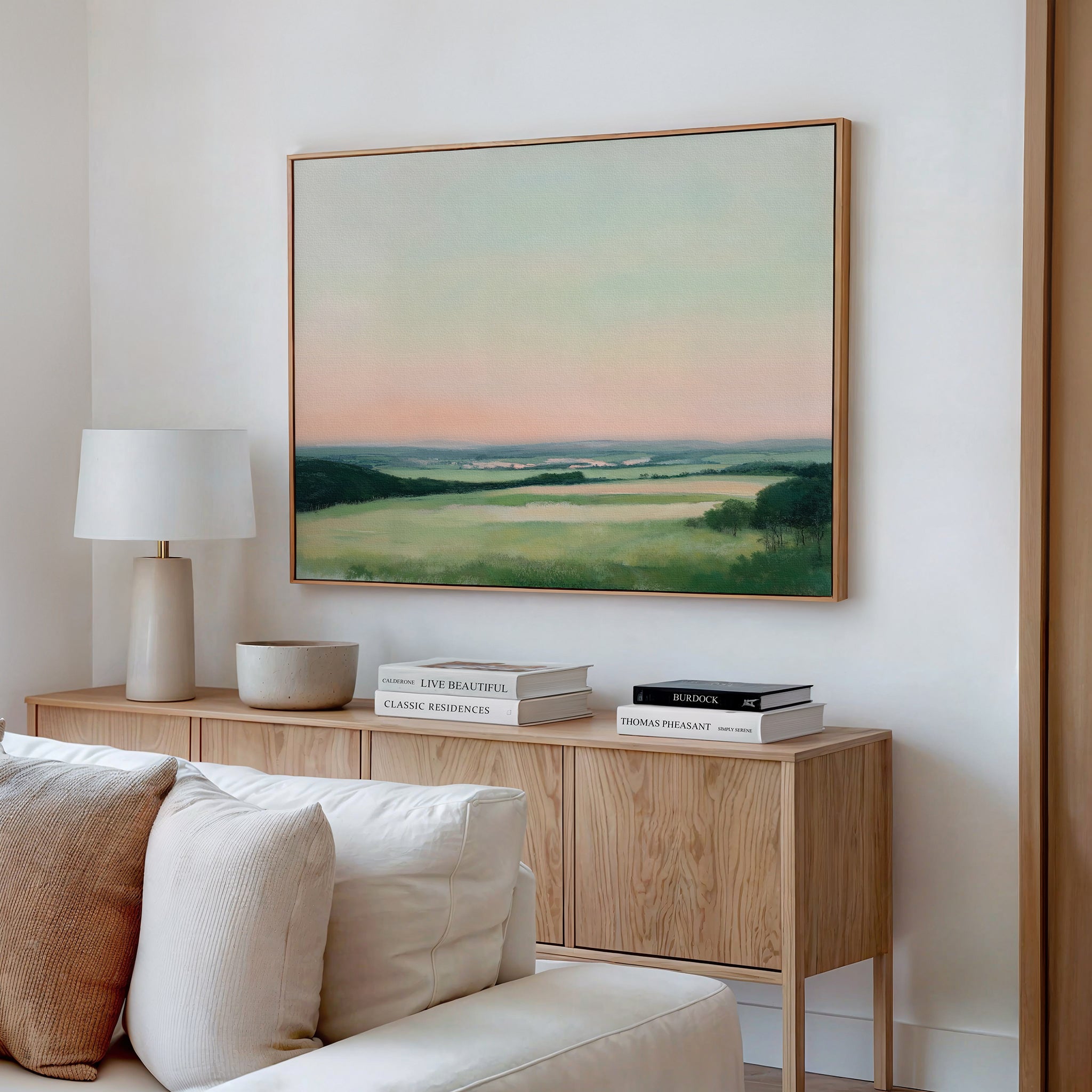 A modern living room features a light wooden console with decorative books, a lamp, and a bowl. Above it hangs the Evening Hues Canvas Art – Horizontal, depicting green fields under a pastel sky. A white sofa sits in the foreground.