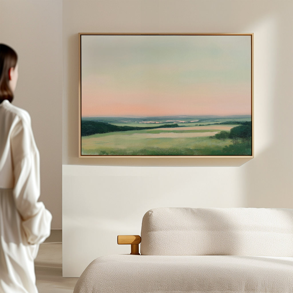A person in a white robe stands in a modern living room, admiring the Evening Hues Canvas Art – Horizontal, featuring soft green fields and a pastel sky above a beige sofa as elegant decor.