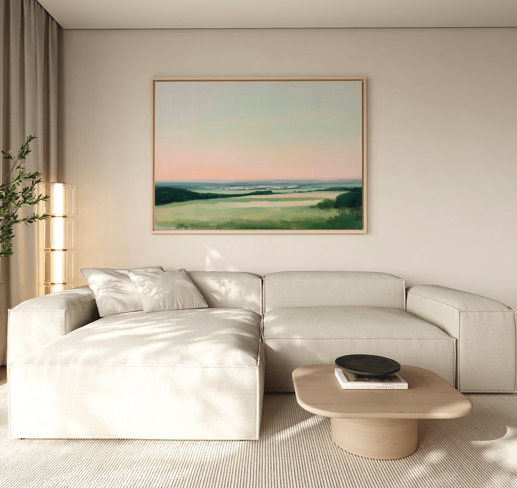 A minimalist living room featuring a light beige sectional, low wood coffee table, and the Evening Hues Canvas Art – Horizontal showing green fields and a pastel sky. A potted plant and tall floor lamp complete the cozy decor.