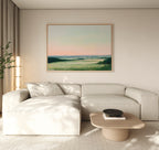 A minimalist living room featuring a light beige sectional, low wood coffee table, and the Evening Hues Canvas Art – Horizontal showing green fields and a pastel sky. A potted plant and tall floor lamp complete the cozy decor.