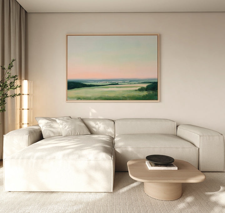 A minimalist living room featuring a light beige sectional, low wood coffee table, and the Evening Hues Canvas Art – Horizontal showing green fields and a pastel sky. A potted plant and tall floor lamp complete the cozy decor.