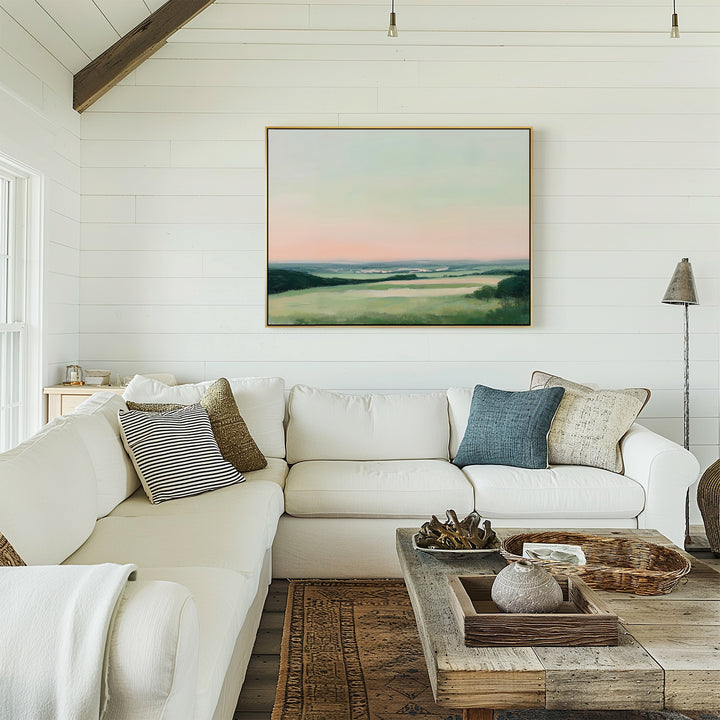 A cozy, bright living room with a white sectional sofa, textured pillows, a rustic wooden coffee table, and the Evening Hues Canvas Art – Horizontal featuring green fields and a pastel sky on the white shiplap wall.