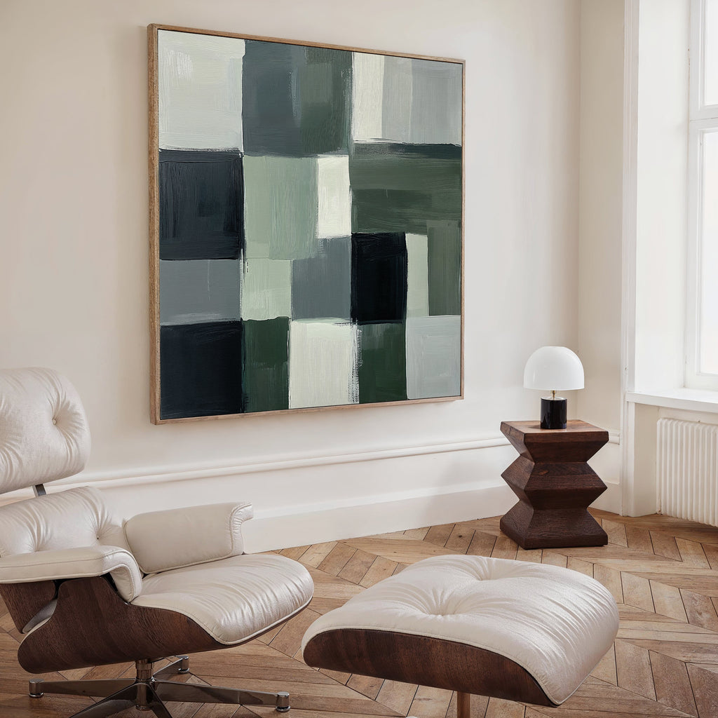 A modern living room featuring a white leather lounge chair and ottoman, a geometric wooden side table with lamp, and the Evergreen Abstract Canvas Art displaying green, gray, and black blocks on the wall.
