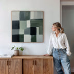 A woman in a white shirt and jeans leans against a wooden cabinet, admiring the Evergreen Abstract Canvas Art with green geometric patterns above her. A potted plant and book on the cabinet add to the modern ambiance.