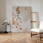 The Faded Flora Canvas Art, featuring blue and brown flowers, leans against a white paneled wall beside a minimalist canvas, black side table with books and candles, and a white cushioned chair on a wooden herringbone floor.