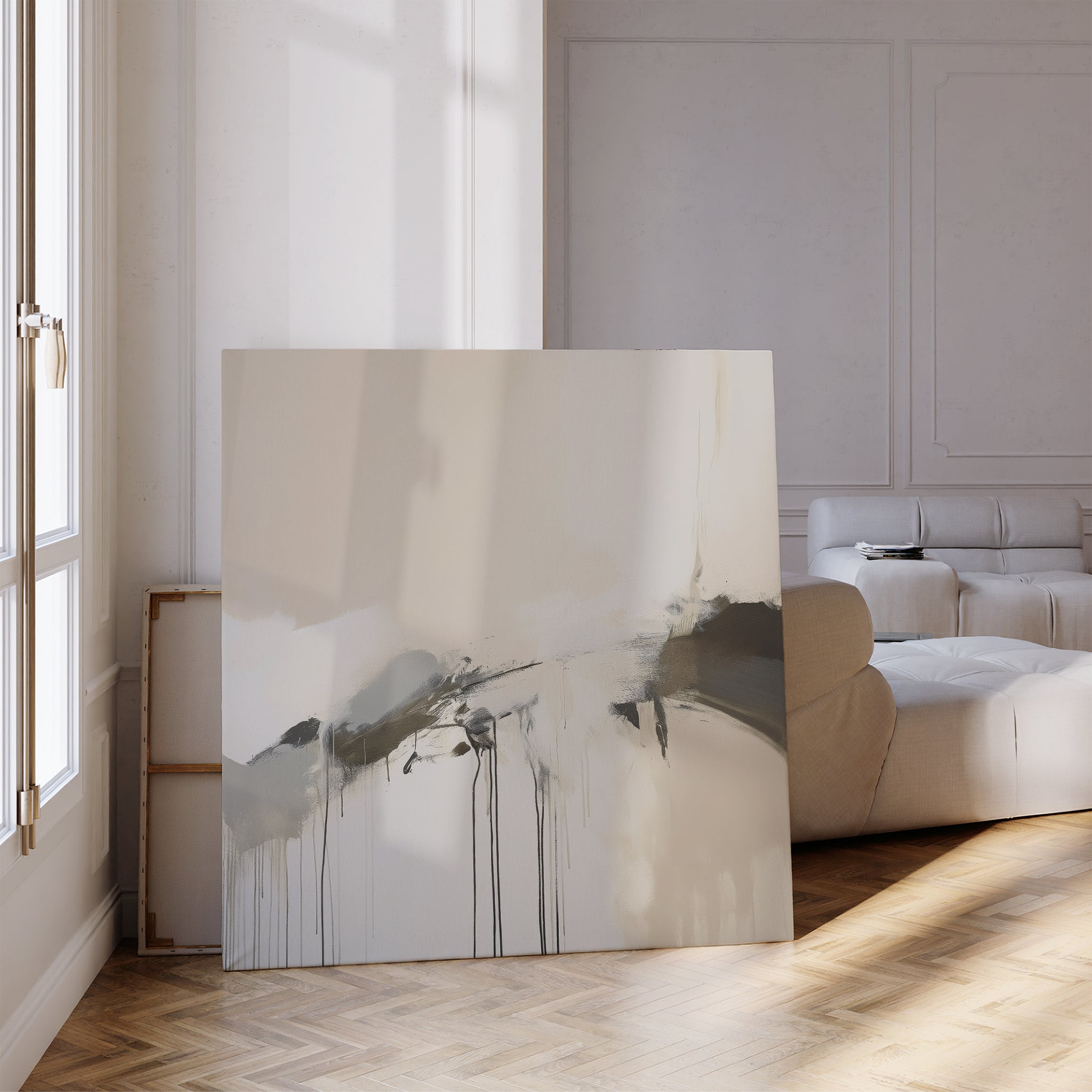 The Faded Memory Canvas Art, featuring grey and black abstract brushstrokes and drips, leans against the wall of a bright minimalist living room with a white sofa, wooden floors, and sunlight streaming through tall windows.