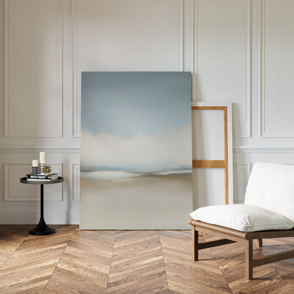 The Faded Silence Canvas Art, featuring minimal softly blurred tones, leans against a white paneled wall beside a cushioned wooden chair and small black table with candles and books atop a herringbone wood floor.